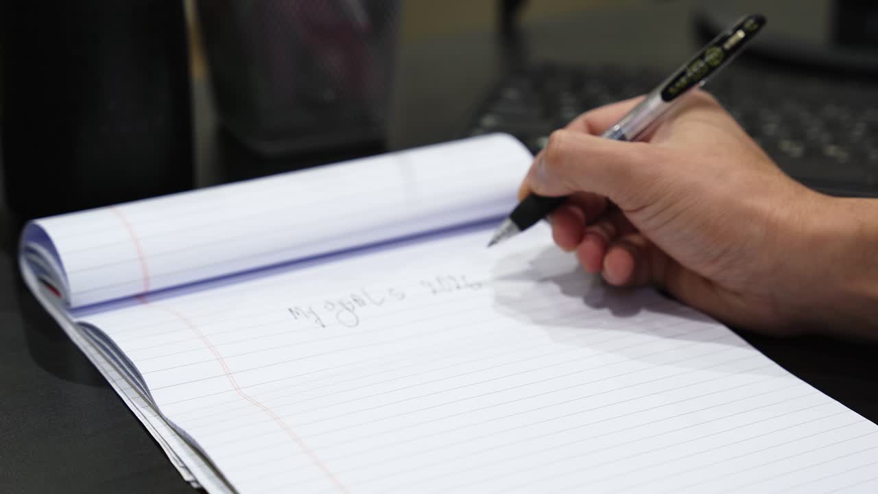 Primo piano di una mano che scrive un elenco numerato di obiettivi in un quaderno a righe su una scrivania, tenendo una penna con tastiera del computer sfocata sullo sfondo sotto l'illuminazione interna dell'ufficio