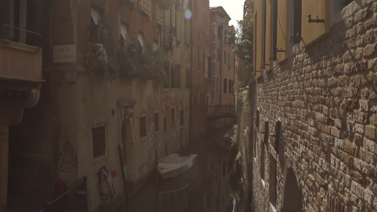 Plano amplo com inclinação para cima da histórica rua do canal aquático Cale Castagna com parede de tijolos, flares solares na lente, Veneza, Itália.