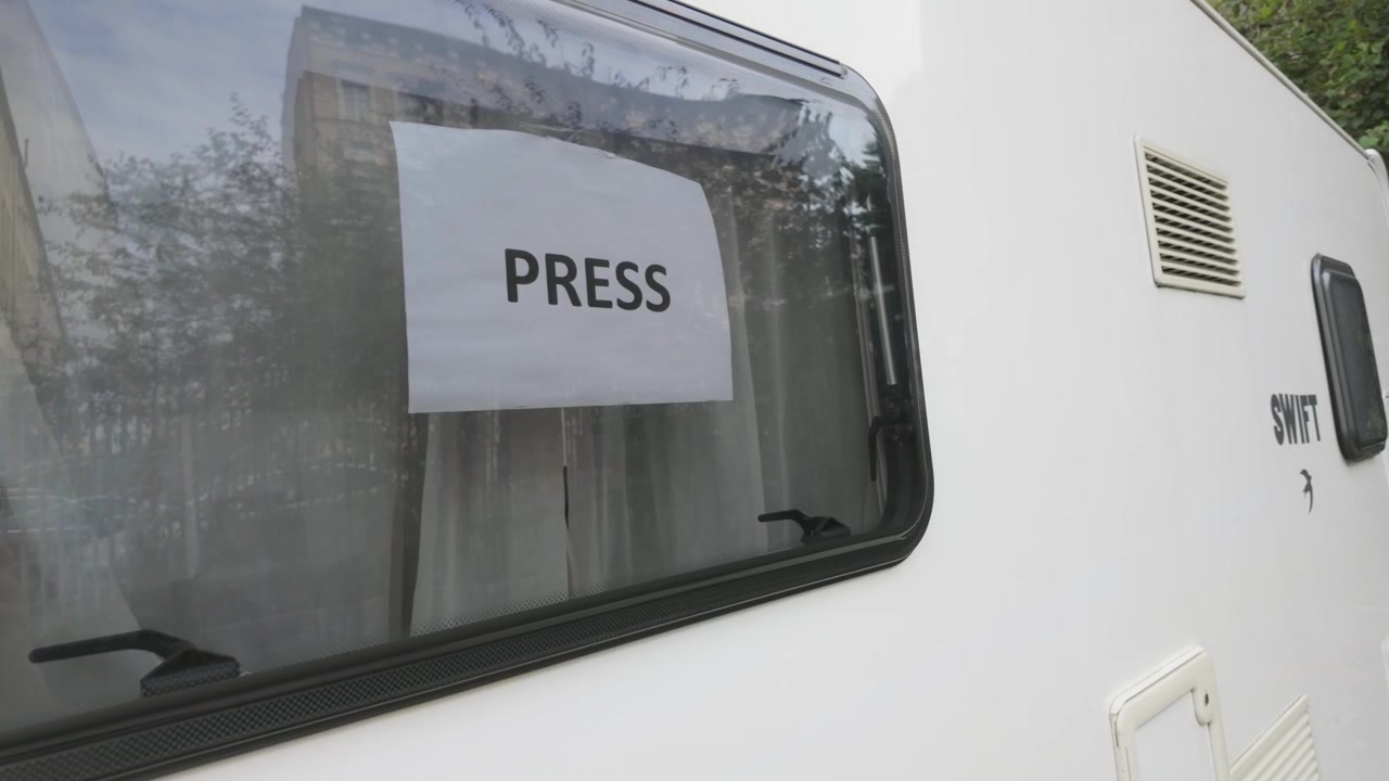 Presse-skilt vist på vinduet af en hvid trailer i et udendørs bymiljø.