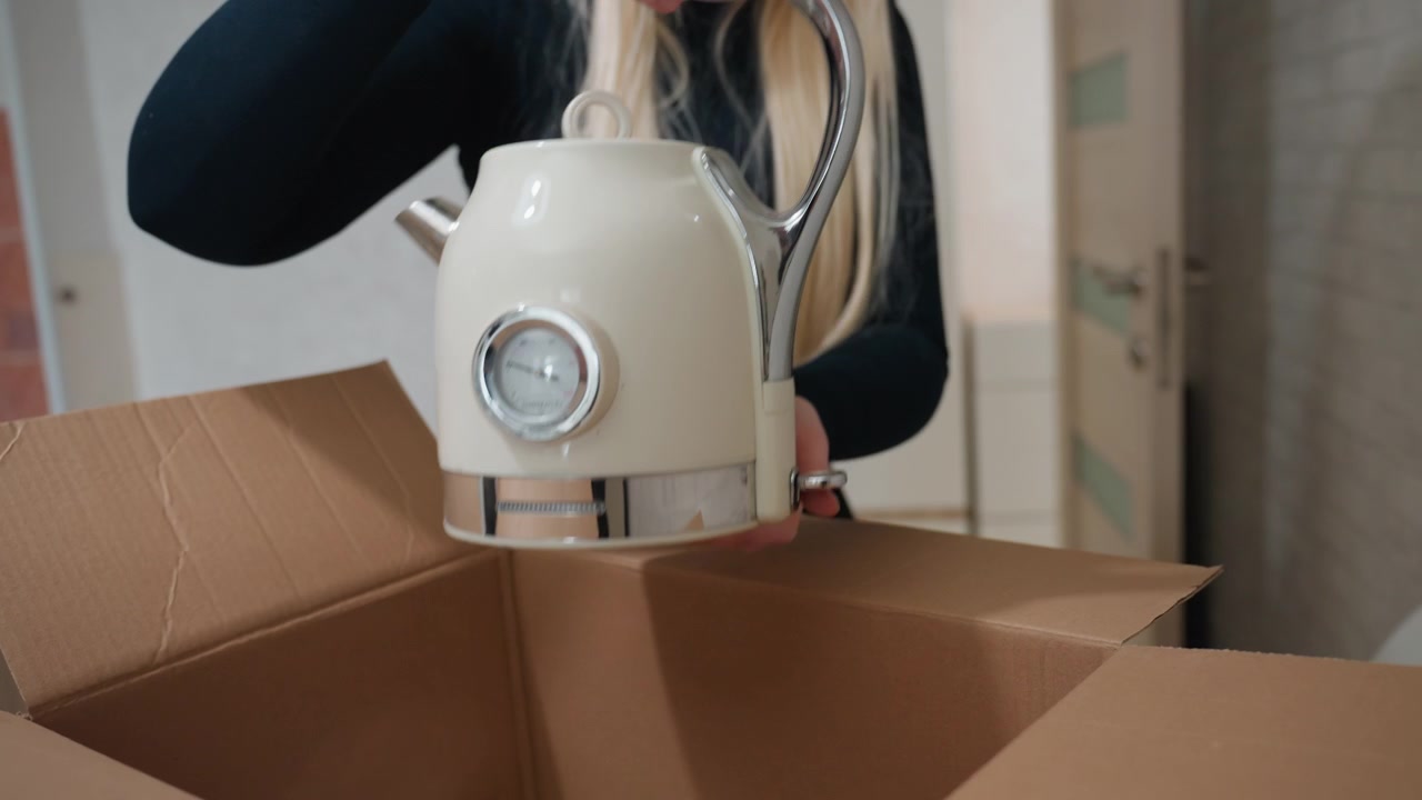 Packing fragile kettle in moving box, closeup of woman hands placing ceramic kettle into cardboard box in apartment, careful handling with padding, organized kitchenware scene, quiet focused mood