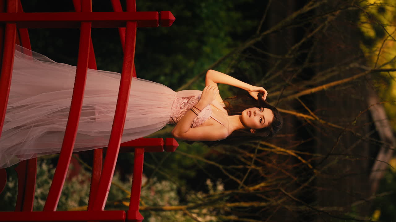 молодая кавказская девушка в длинном розовом платье модели на мосту ботанического сада, вертикальное видео при дневном свете, бледно-белая осень, играя с волосами