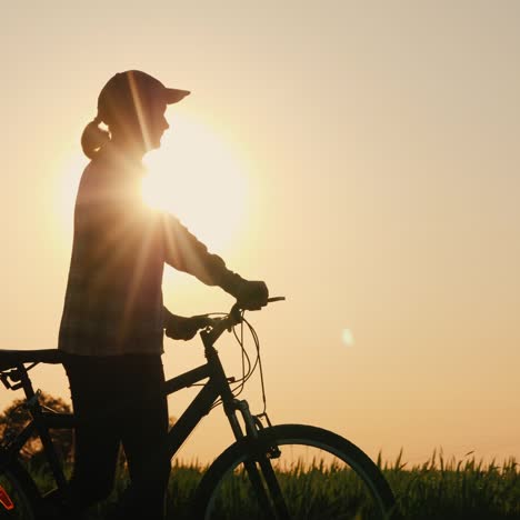 Die-Silhouette-Einer-Frau,-Die-Bei-Sonnenuntergang-Ein-Fahrrad-Führt-Und-Die-Natur-Bewundert