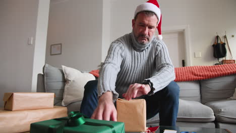 senior man wrapping christmas gifts at home, wearing santa hat