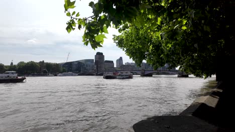 Cityscape-view-of-the-Thames-river-featuring-boats-and-buildings,-with-trees-in-the-forefront,-capturing-the-essence-of-a-bustling-urban-environment-on-a-calm-day