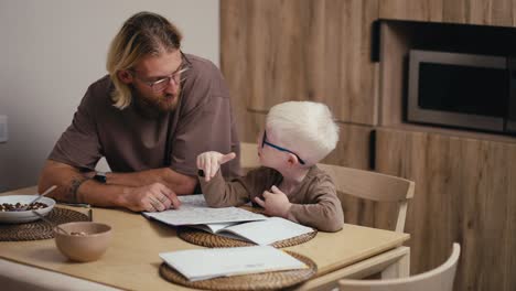 Happy-blond-man-with-a-beard-and-glasses-helps-his-little-albino-son-with-short-white-hair-in-blue-glasses-solve-his-homework-and-write-the-correct-answers-in-the-questions-while-preparing-his-homework-at-home-in-the-kitchen