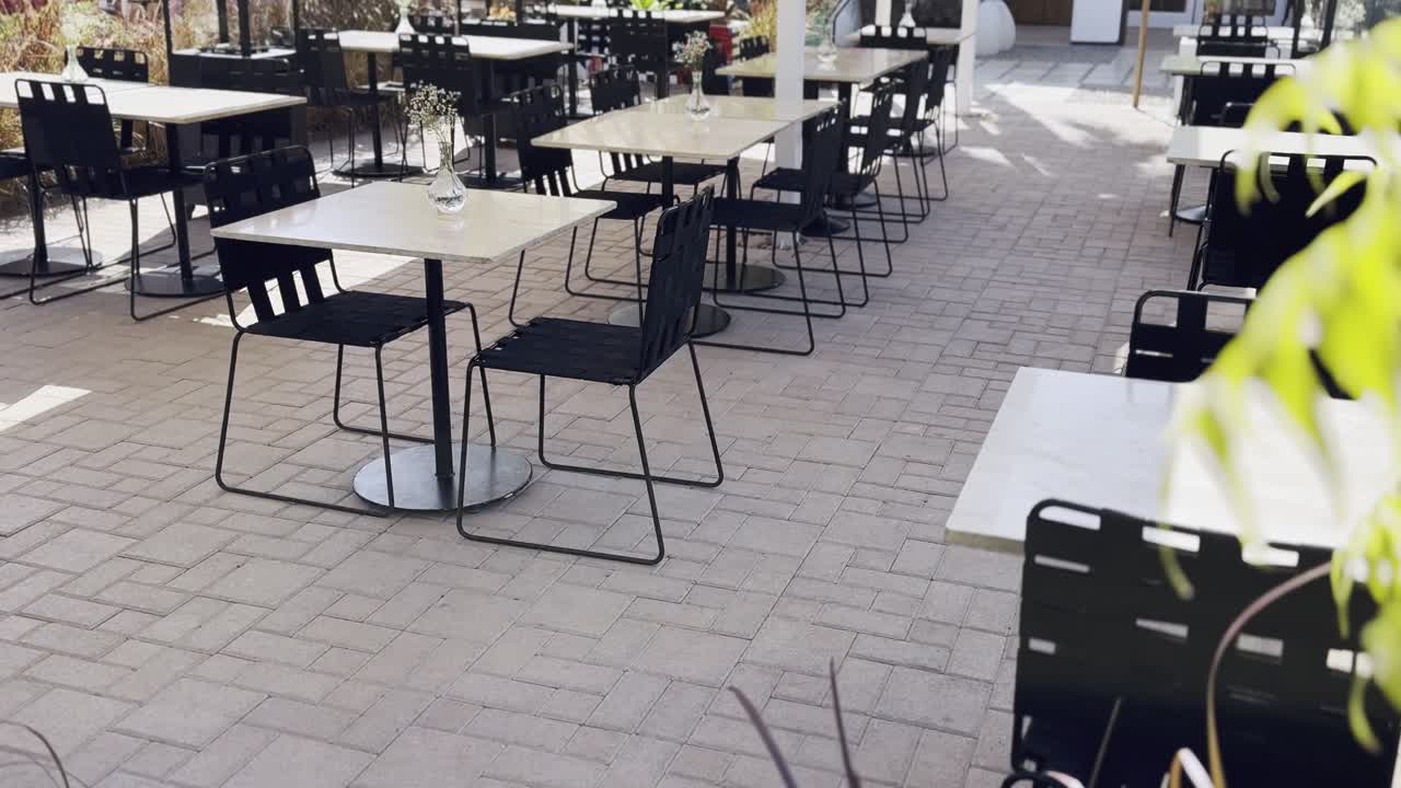 Una terrazza di un bar all'aperto, inondata di sole, con tavoli bianchi moderni, sedie nere e piante verdeggianti su un patio pavimentato durante una giornata luminosa