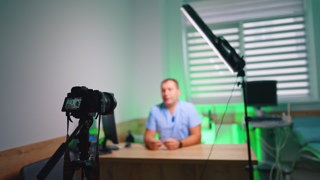Allestimento per vlog clinico con medico, telecamera e luce. Ampia configurazione clinica con telecamera su treppiede, luce a tubo LED, scrivania e medico sfocato sullo sfondo