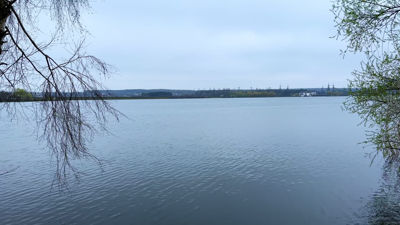At the bank of calm river on gloomy spring day. Some branches hang over the water. Other side of the river at backdrop.