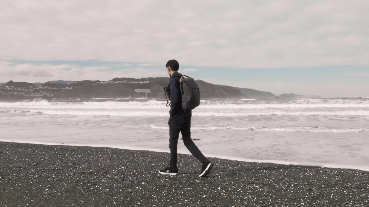 Seorang Pria Berjalan di Pantai Berbatu dengan Ombak Menerjang di Teluk Lyall, Wellington, Selandia Baru. Rekaman Gerak Lambat.