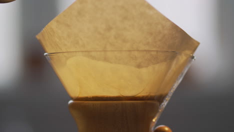 close up view of kettle pouring hot water into large glass chemex coffee maker over fresh, ground coffee beans in the morning