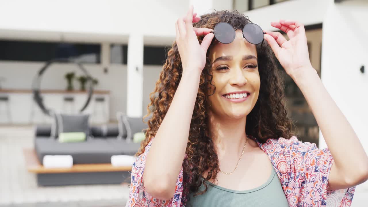 video de una feliz mujer biracial levantando gafas de sol y sonriendo al sol en un complejo vacacional