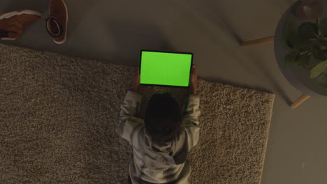 overhead shot of young boy lying on rug at home at home playing games or streaming onto green screen digital tablet at night 1