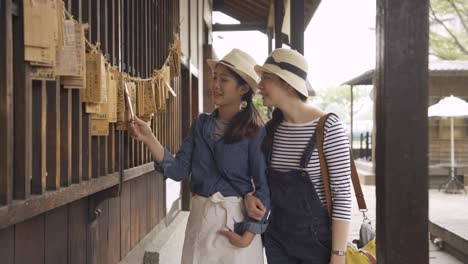 two asian girls tourists travel tokyo japan walking in temple corridor. young women traveler standing in hallway hands holding sharing hope board on wall curious reading letter discussing laughing