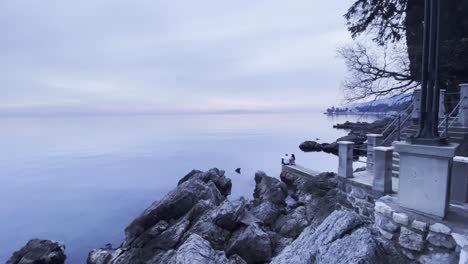 couple sitting on sea shore with island in distance fish eye wide lovran, opatija, croatia