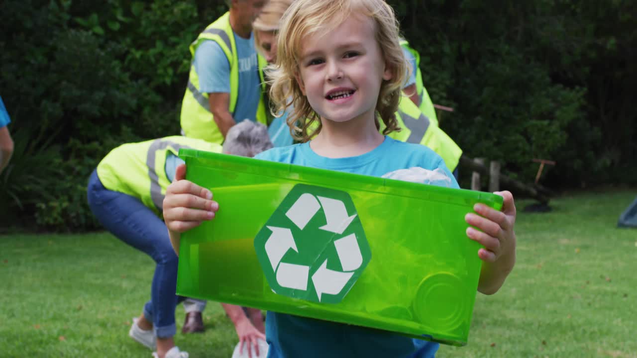 niño caucásico sonriente sosteniendo una caja de reciclaje recogiendo basura con voluntarios en el campo