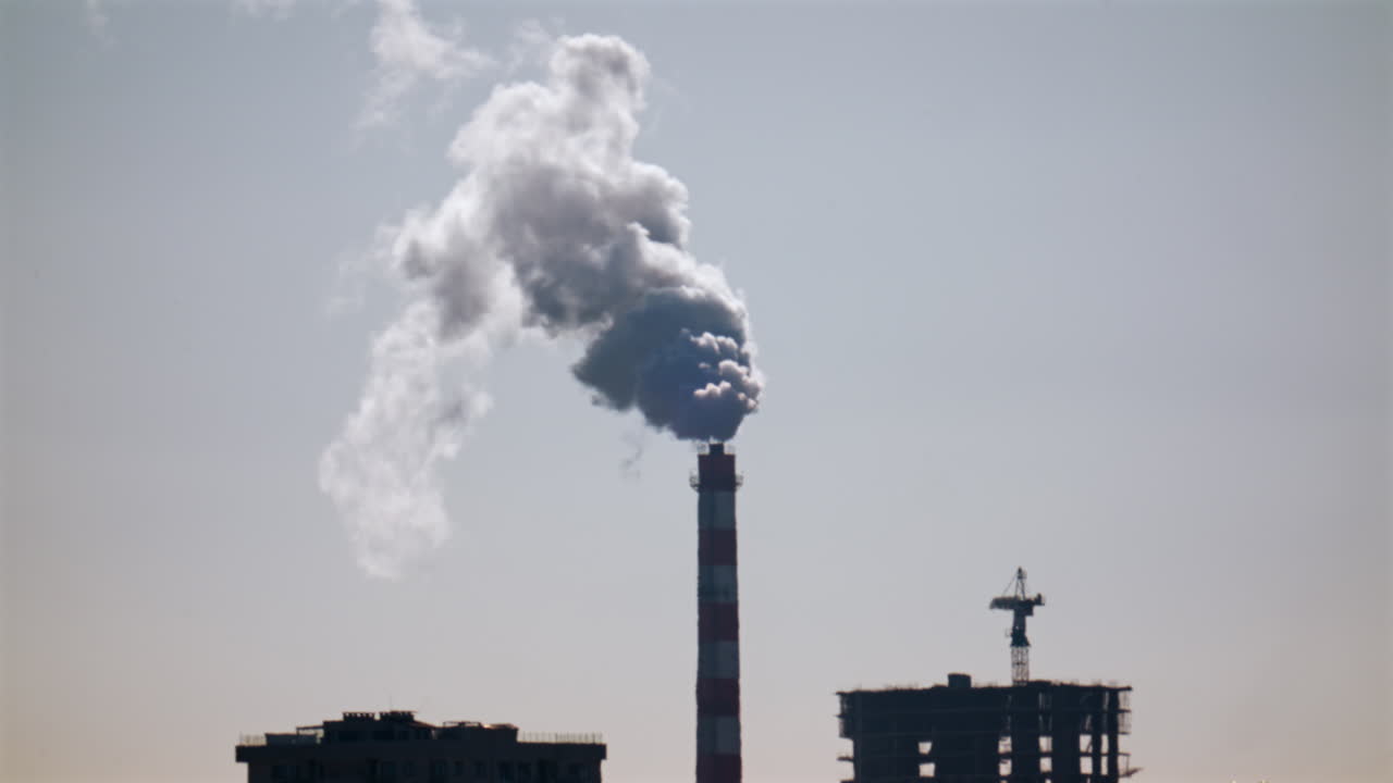 Ciminiera industriale che rilascia fumo denso nel cielo sopra un cantiere edile cittadino.