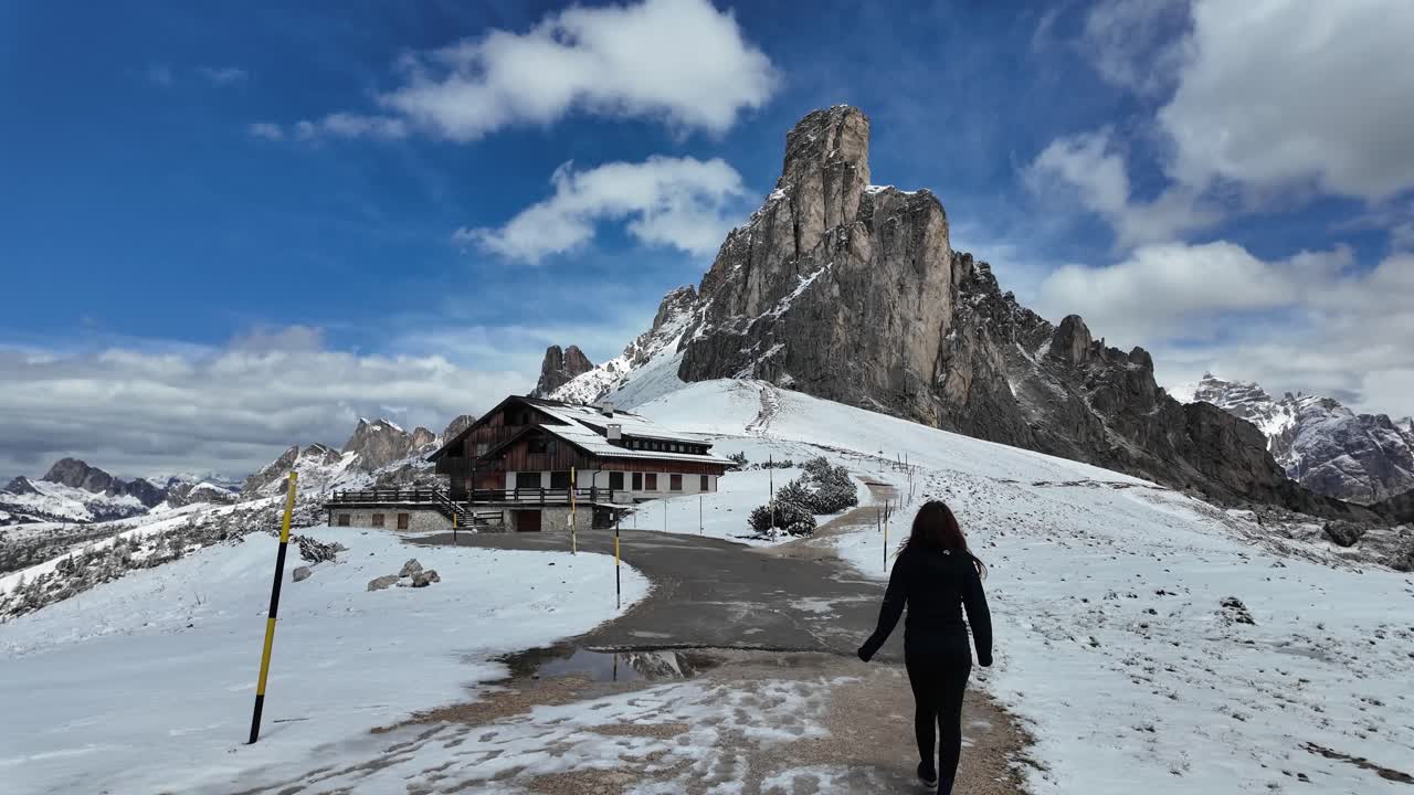 A video of a girl walking in Passo Giau, Italian Dolomites, with snow-covered peaks and a rustic cabin in the background