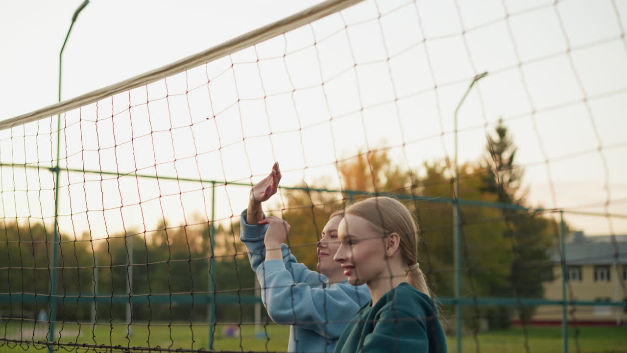 Teammates jumping together with joy, sending volleyball back across the net, both in green and blue hoodies, smiling and celebrating successful play in outdoor volleyball match