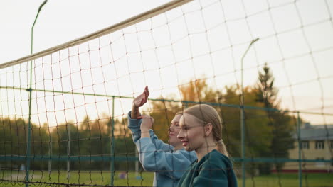 teammates jumping together with joy, sending volleyball back across the net, both in green and blue hoodies, smiling and celebrating successful play in outdoor volleyball match