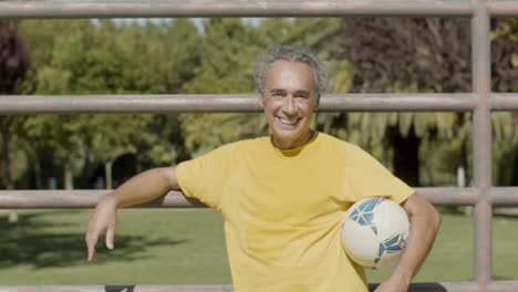 vista frontal de un feliz jugador de fútbol senior sosteniendo la pelota, levantando el brazo y mirando la cámara