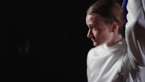 a young female fencer in a white uniform, holding a fencing mask and sword, looks directly at the camera with a serious expression.