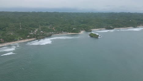 Una-Exuberante-Playa-Indonesia-Con-Olas-Y-Un-Islote-Solitario,-Día-Nublado,-Vista-Aérea