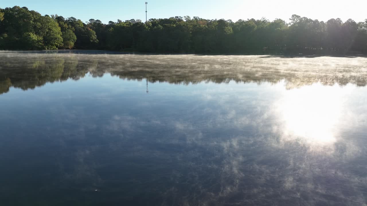 Morning fog drifts across a calm lake as sunlight reflects off the still water