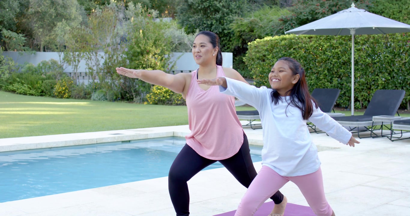 Người mẹ châu Á thực hiện tư thế lunge, cô bé Mỹ gốc Phi bắt chước theo, cả hai tập yoga trên thảm tím cạnh hồ bơi.