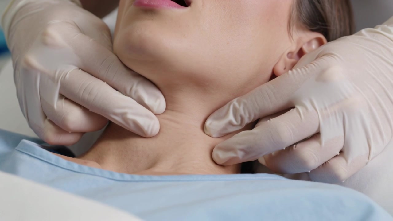 Clinico guantato che si avvicina e palpa il collo del paziente sul lettino da visita, valutando il collo, con camice blu.