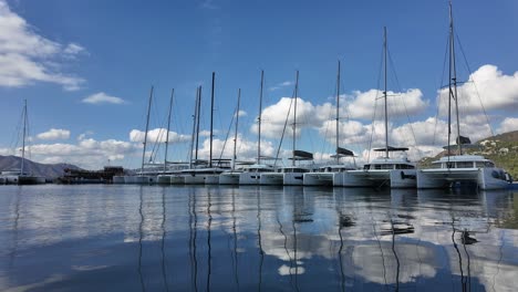 nine catamarans docked side by side at the marina on a sunny day,white clouds moving above their poles and reflections on the blue calm sea.