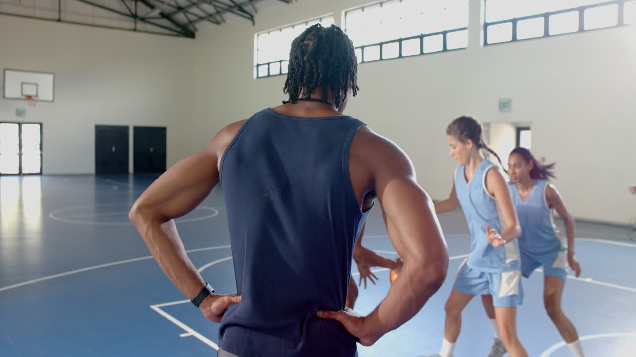 Coach observing female basketball players practicing dribbling and defense in gym