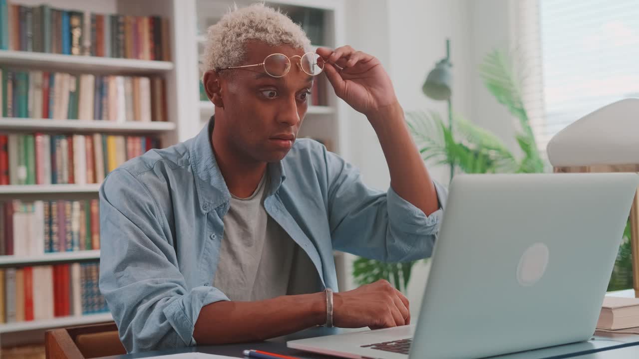 Young african american man raises glasses and opening mouth looks at laptop