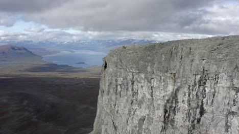 A-dramatic-aerial-drone-shot-of-some-people-stood-on-the-top-of-a-huge-cliff-looking-over-a-vast-mountainous-landscape