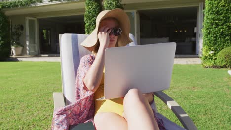 caucasian woman using laptop while sunbathing on a deck chair in the garden