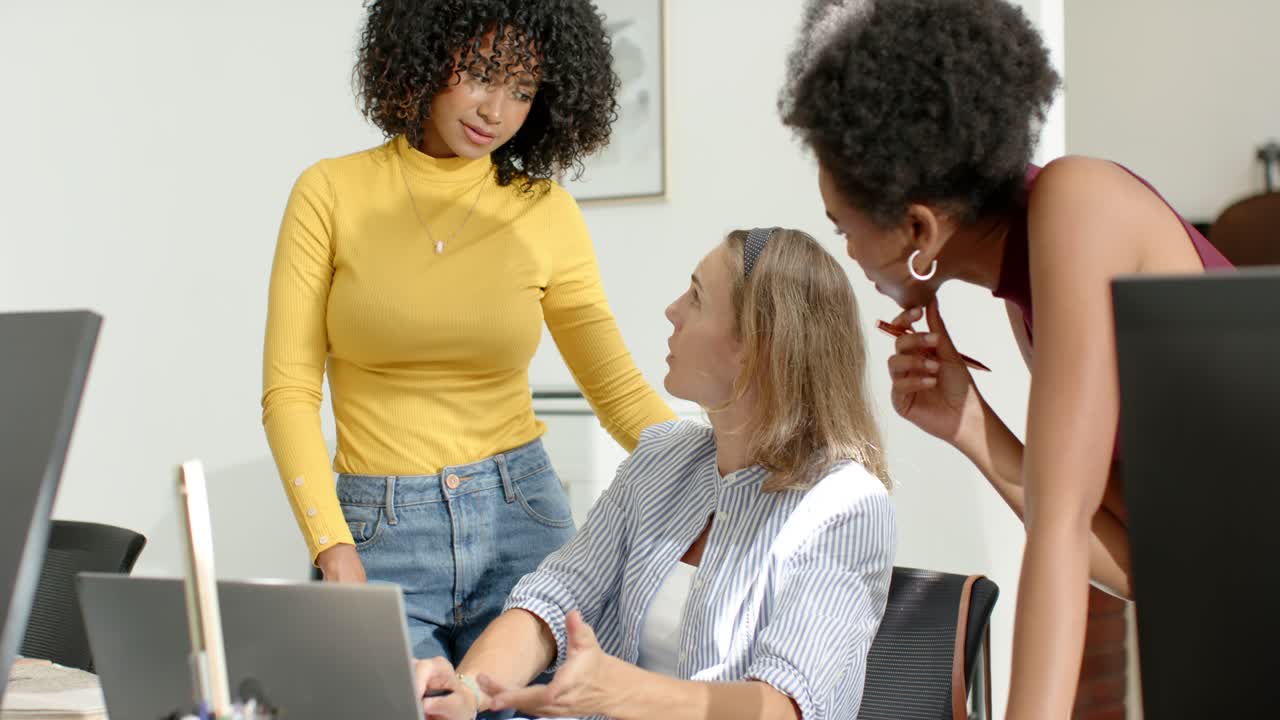 Colegas de trabalho diversas inclinadas na mesa revisando o laptop enquanto uma mulher sentada digita e explica