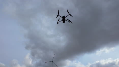 Quadrocopter-Drohne-Schwebt-Im-Himmel-Mit-Wolken-Und-Einer-Windturbine-Im-Hintergrund