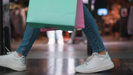 side leg view of someone in jeans and white canvas sneakers carrying mint and pink shopping bags in their right hand while walking past a clothing store in a brightly lit shopping mall