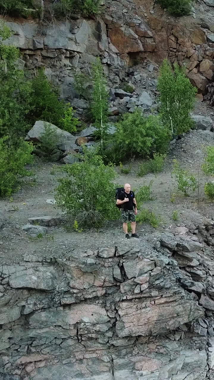 Young tourist man standing on steep slope with backpack over the water. Camper observing beautiful view of canyon with green trees and bushes and their reflection in the water. Aerial view Vertical video