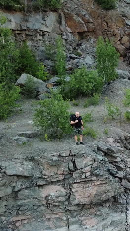 young tourist man standing on steep slope with backpack over the water. camper observing beautiful view of canyon with green trees and bushes and their reflection in the water. aerial view vertical video