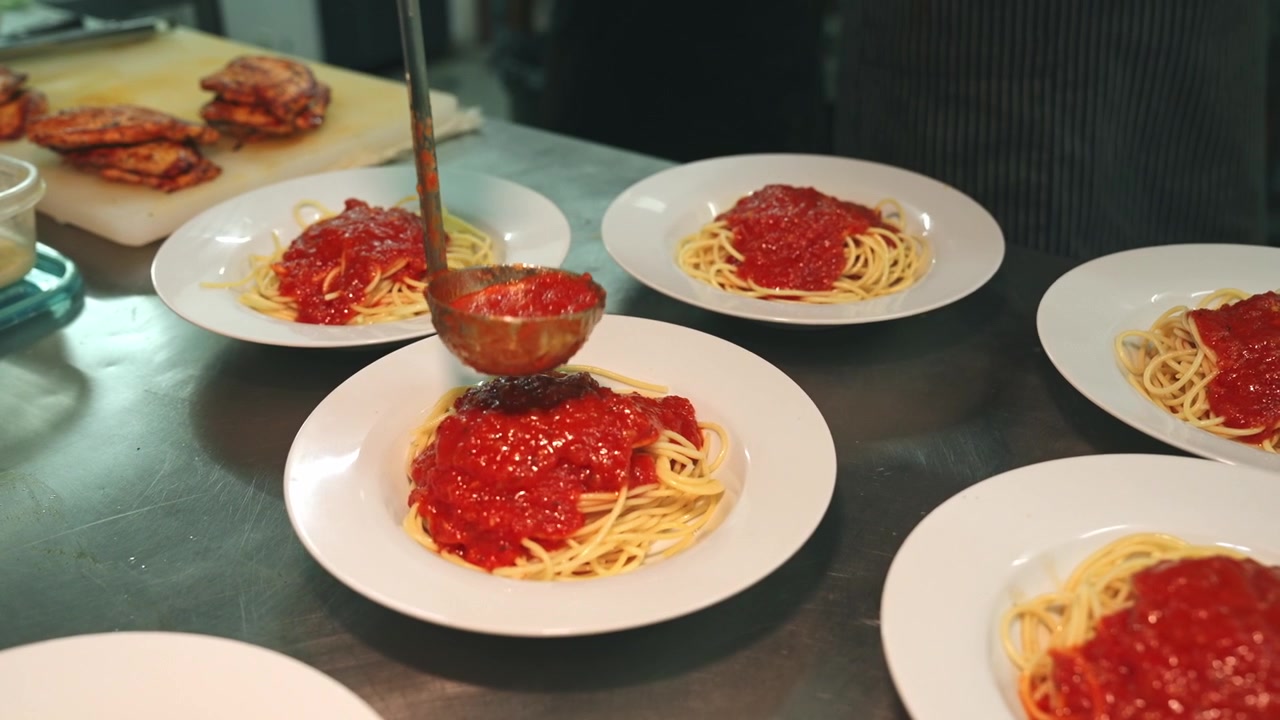 Inquadratura Media in Movimento di un Bancone di Cucina Commerciale dove uno Chef con Guanti Neri Versa Sistematicamente con un Mestolo Salsa di Pomodoro Calda su Più Piatti di Spaghetti