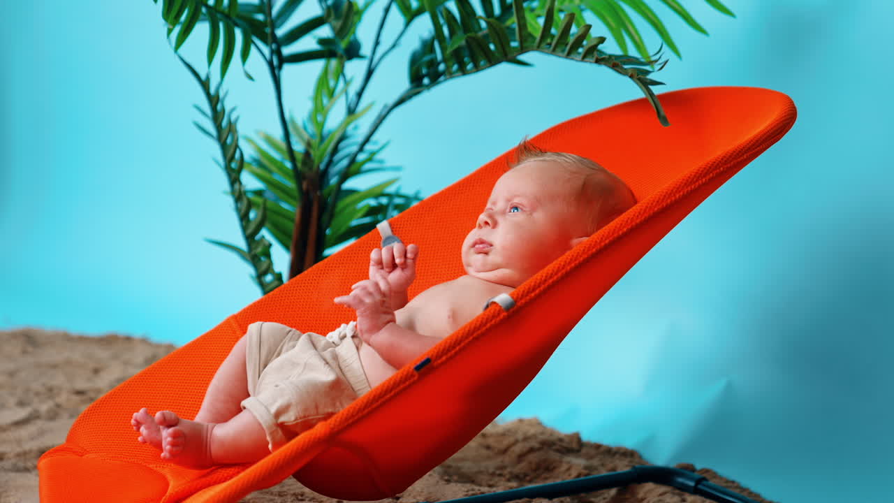 Lovely blond Caucasian baby in beige shorts lies resting in the baby chair. Sand and plant in the pot at backdrop.