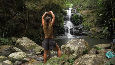 mujer despreocupada levantando ambas manos con una cascada prístina y un río en el fondo - parque nacional lamington en la costa dorada, australia