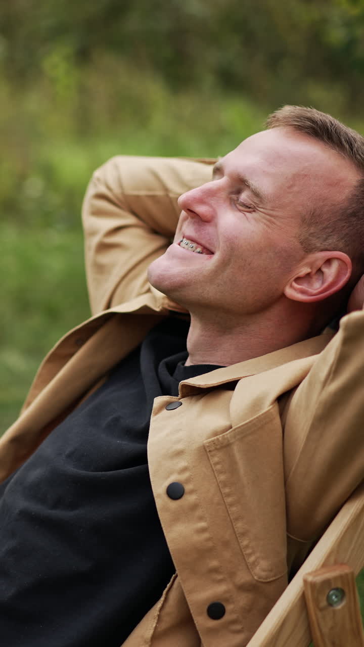 Happy man relaxing and rejoicing sitting in the garden chair outdoors. Caucasian male finished remote work on his laptop and sits smiling to the camera. Vertical video