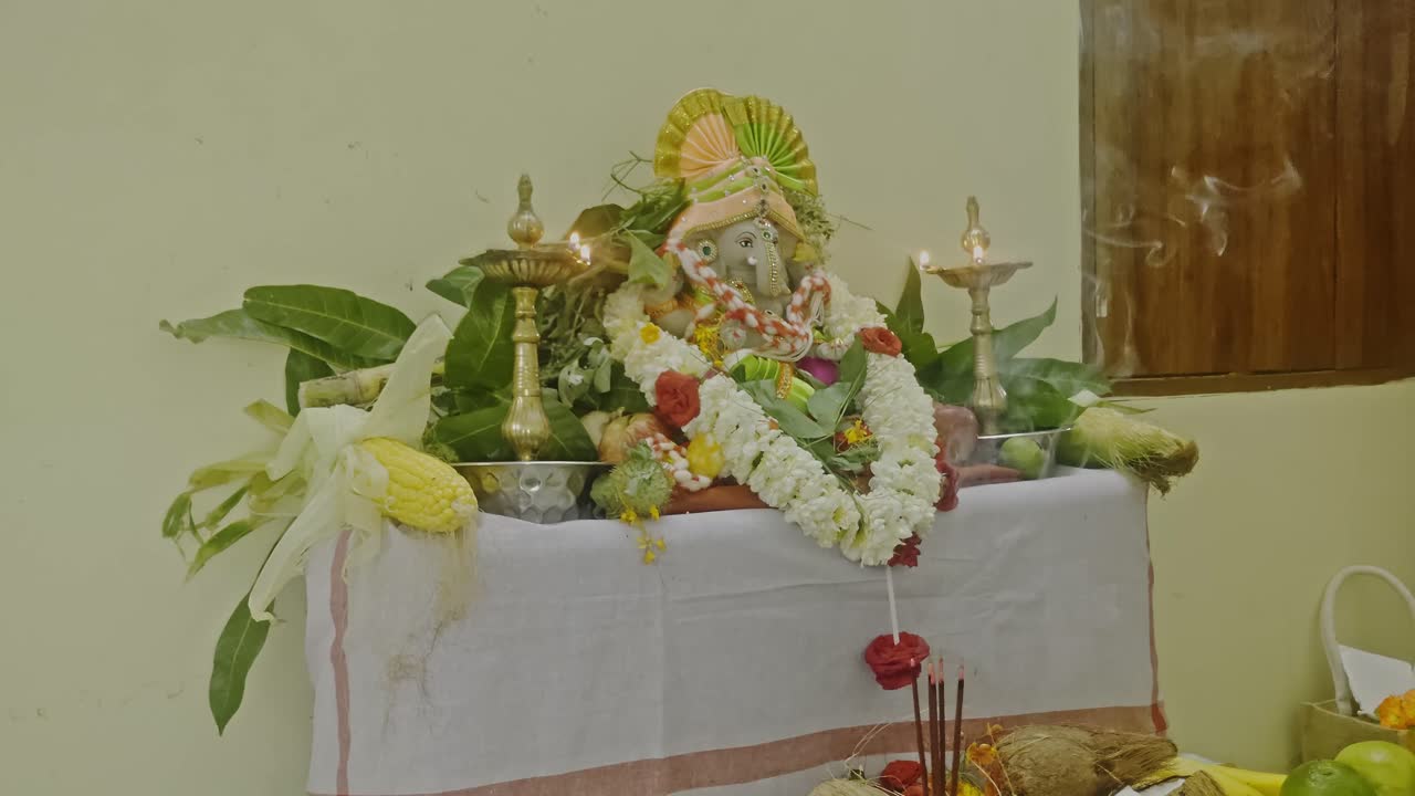 Lord Ganesh posizionato su un altare tradizionale, adornato con ghirlande, fiori, frutta e lampade a olio accese. Scatto interno, scatto stabile, 4K