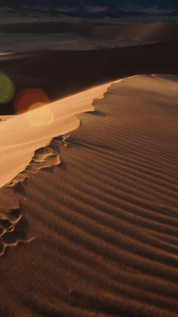 Video verticale: Telecamera che si muove lungo la cresta di una duna all'alba per rivelare increspature, macchie scure e un bagliore dell'obiettivo.