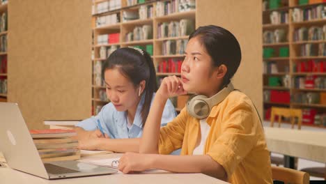 no idea asian woman student with headphones thinking so hard to write into the notebook while sitting with her classmate on a table with a laptop in the library