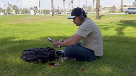 a man is sitting on the grass wearing headphones and he is reading something on the tablet.