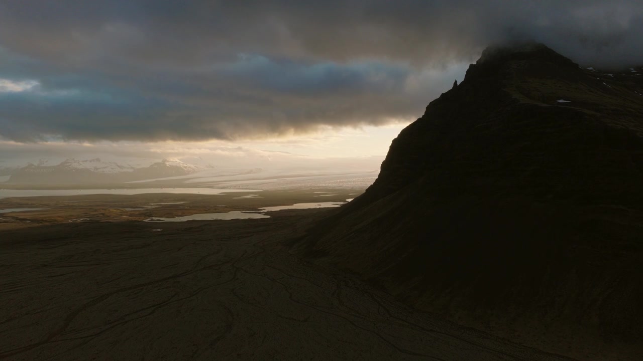 Herdisarvikurfjall rises above the volcanic coastal plain of Herdisarvik, Olfus, Iceland, with dark lava slopes and the Atlantic coastline fading through golden haze, drone forward flyover.