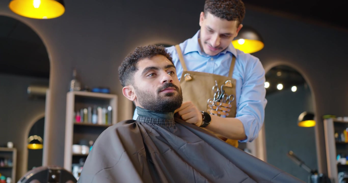 Afroamerikansk barber lægger en sort kappe på kunden, fæstner kraven og forbereder klipningen i barbersalonen.