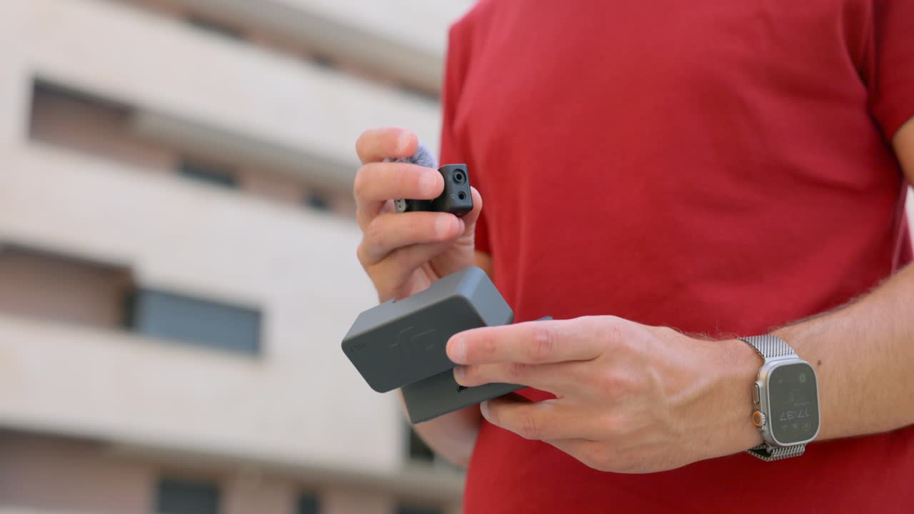Primo piano di un uomo che tiene in mano un set di microfoni wireless e la custodia di ricarica all'aperto, mentre prepara l'attrezzatura audio per le riprese.